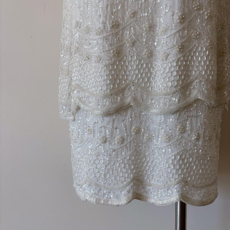 May include: A white, beaded dress with a layered design. The dress features intricate patterns of sequins and beads, creating a textured and shimmering effect. The dress is displayed on a silver clothing rack.