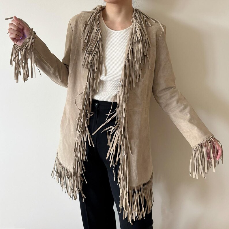 May include: A tan suede jacket with long sleeves and a fringed design. The jacket features fringe along the collar, front, and sleeves. The jacket is open, revealing a white top and black pants.