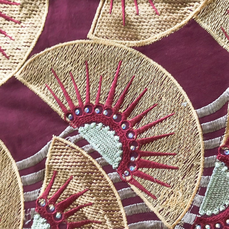 May include: Close-up of a fabric with an embroidered fan design. The fans are gold with red accents and embellished with rhinestones. The background is a deep burgundy color, creating a striking contrast.