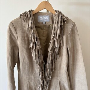 May include: A tan suede jacket with fringe detailing. The jacket features a notched collar and a button closure. The brand name "Wilsons Leather Maxima" is visible on a tag inside the jacket. The jacket is hanging on a wooden hanger.