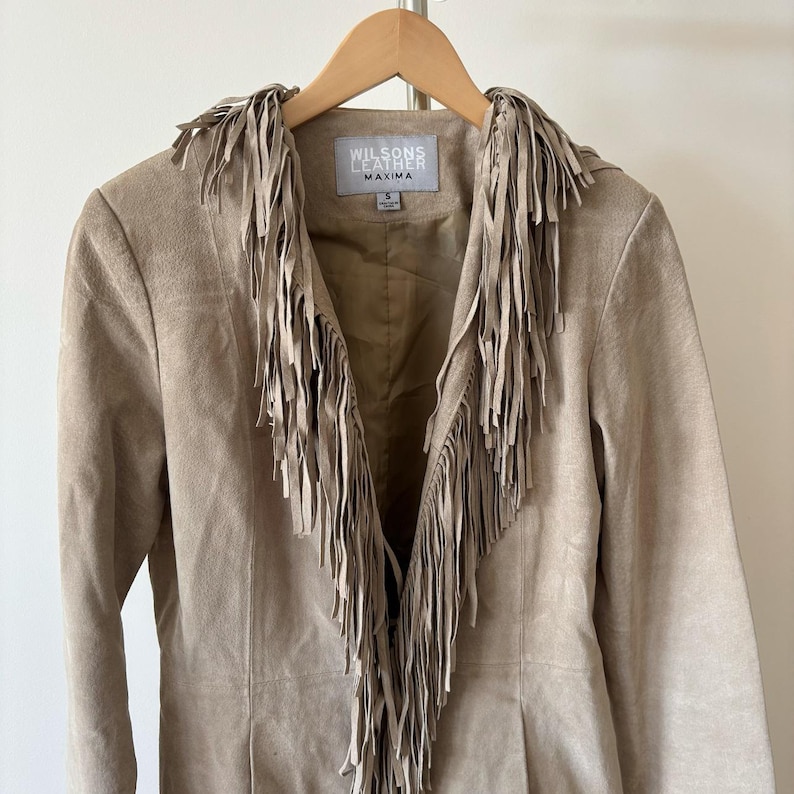 May include: A tan suede jacket with fringe detailing. The jacket features a notched collar and a button closure. The brand name "Wilsons Leather Maxima" is visible on a tag inside the jacket. The jacket is hanging on a wooden hanger.