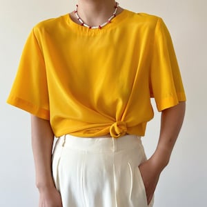 Puede incluir: Una blusa amarilla brillante de manga corta con un nudo en la parte delantera. La blusa est&aacute; hecha de un material sedoso y tiene un escote redondo. La modelo lleva un collar blanco con detalles rojos.