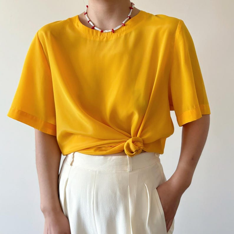 Puede incluir: Una blusa amarilla brillante de manga corta con un nudo en la parte delantera. La blusa est&aacute; hecha de un material sedoso y tiene un escote redondo. La modelo lleva un collar blanco con detalles rojos.