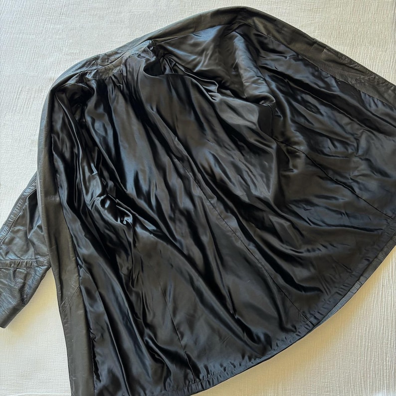 May include: A black leather jacket, open to reveal a black satin lining. The jacket is laid out on a white surface, showcasing the interior construction and design. The leather appears worn, adding character.