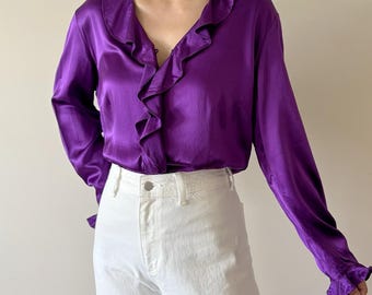May include: A vibrant purple long-sleeve blouse featuring a deep V-neckline with cascading ruffles along the front opening and cuffs. The garment is made of a smooth, satin-like fabric and is paired with plain white trousers with a visible button.