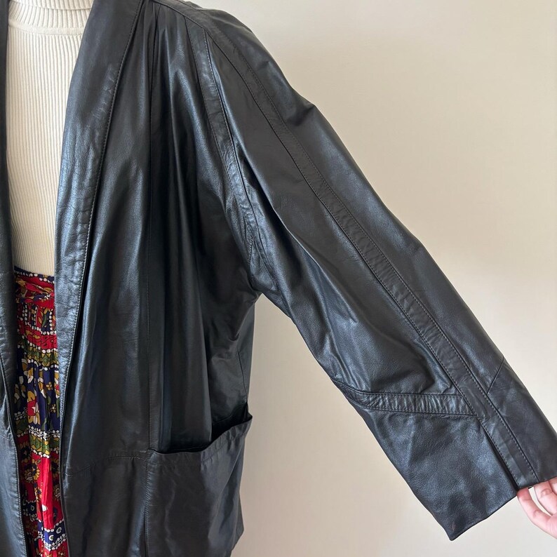 May include: Black leather jacket with a shawl collar and long sleeves. The jacket has a relaxed fit and a pocket on the front. The sleeves have a geometric design near the elbow. The jacket is worn over a white turtleneck and a patterned skirt.