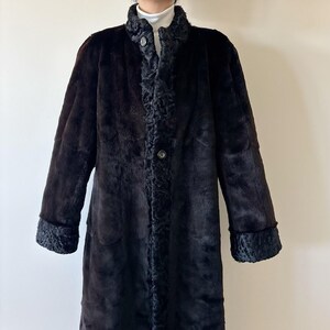May include: A long, dark brown coat featuring a smooth, plush texture throughout the body. The garment includes a textured, curly-patterned trim along the front placket, collar, and sleeve cuffs, secured by visible dark buttons down the center.