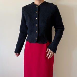 Puede incluir: Chaqueta negra texturizada con cuello redondo y botones dorados. La chaqueta tiene mangas largas y dobladillo festoneado. Combinada con una falda roja. Un estilo cl&aacute;sico y vintage.