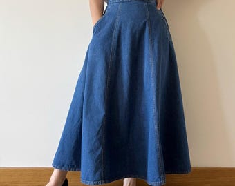 May include: A mid-blue denim A-line skirt. The skirt has a high waist, pockets, and vertical seams. The midi-length skirt is made from a sturdy denim fabric. The skirt's design is classic and versatile.
