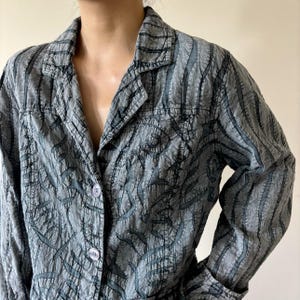May include: A gray button-down shirt with a textured, patterned design. The shirt features a collared neckline, long sleeves, and a button closure. The pattern includes dark gray, abstract shapes and vertical lines. The fabric appears to have a crinkled texture.