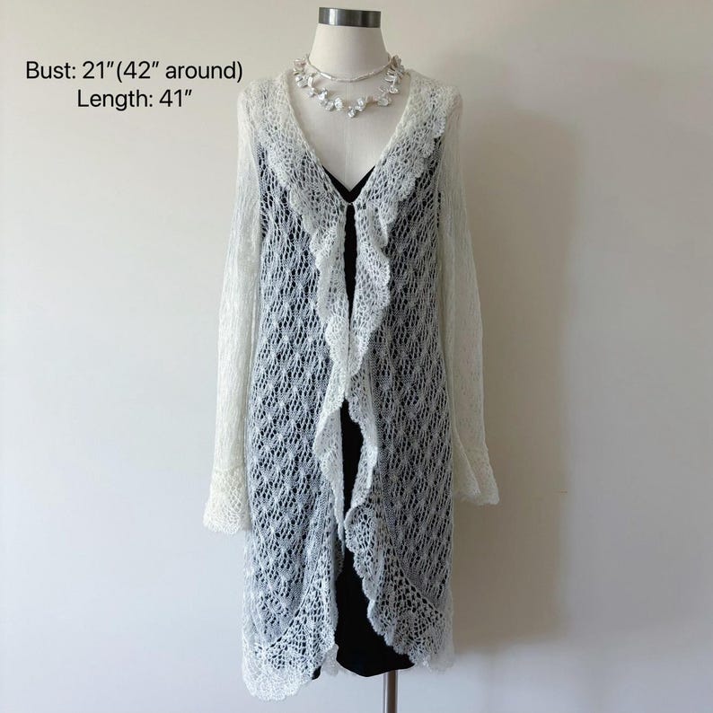 May include: A white, open-front, long-sleeved cardigan with a delicate, open-weave design. The cardigan features a ruffled trim along the front and sleeves, and is 41 inches long. A white floral necklace is also displayed.
