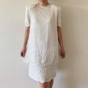 May include: A white, two-piece dress with short sleeves and a round neckline. The dress is covered in white beads and sequins, creating a textured, shimmering effect. The skirt has a scalloped hemline.