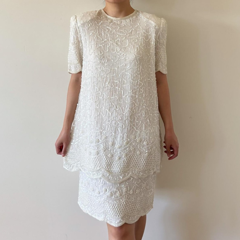 May include: A white, two-piece dress with short sleeves and a round neckline. The dress is covered in white beads and sequins, creating a textured, shimmering effect. The skirt has a scalloped hemline.