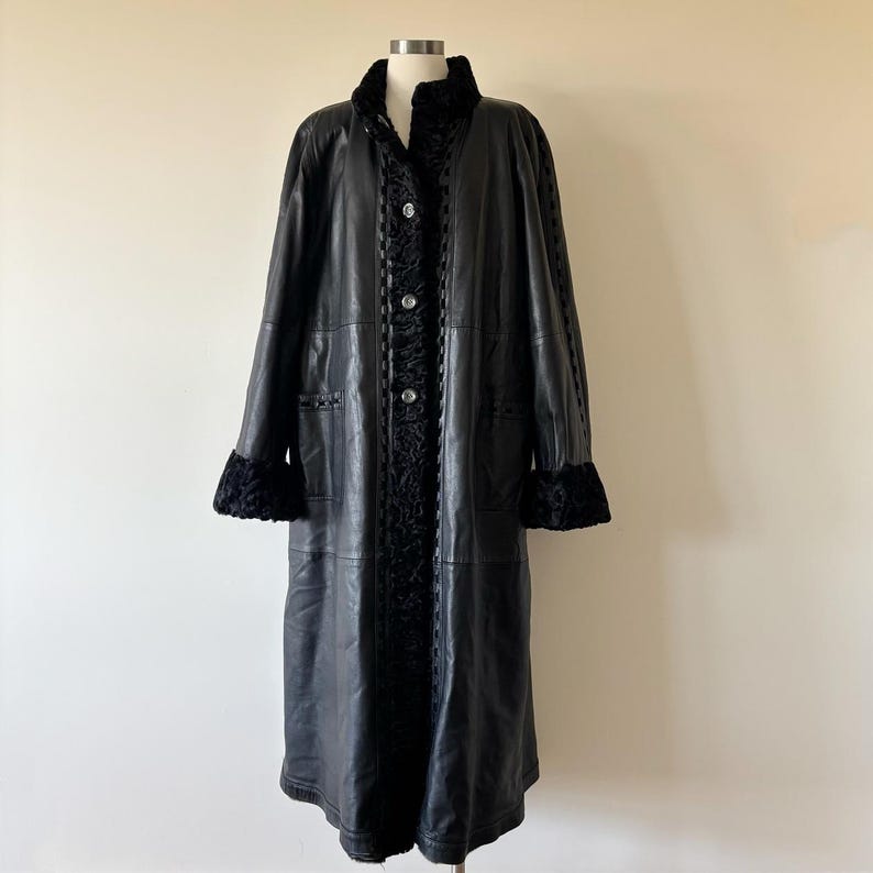 May include: A long black leather coat featuring a textured black fur-like collar, cuffs, and a central vertical panel. The coat includes a front button closure with three visible buttons and a decorative braided trim along the front opening.