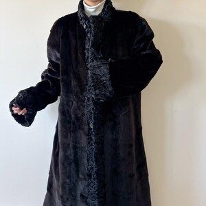 May include: A long, black coat featuring a textured, curly faux fur trim along the front placket, collar, and sleeve cuffs. The main body of the coat has a smooth, dark finish, and it is fastened with a single visible button at the center front.