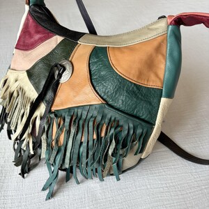 May include: A patchwork leather shoulder bag with a variety of colors including green, tan, cream, and burgundy. The bag features fringe detailing and a silver-toned metal emblem. The strap is black.