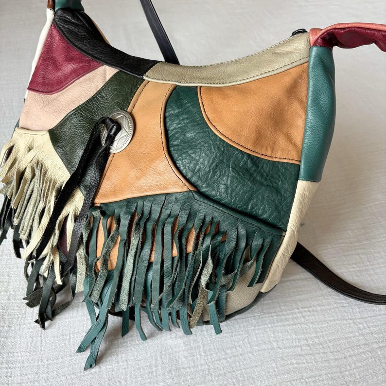 May include: A patchwork leather shoulder bag with a variety of colors including green, tan, cream, and burgundy. The bag features fringe detailing and a silver-toned metal emblem. The strap is black.