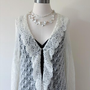 May include: A white, open-front, lace-knit cardigan with a scalloped edge, displayed on a mannequin. A black top is visible underneath. Two pearl necklaces are draped around the mannequin's neck, adding a touch of elegance to the ensemble.