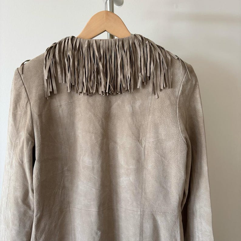 May include: A tan suede jacket with a fringed collar, hanging on a wooden hanger. The jacket has a classic design with vertical seams and a neutral color palette, suitable for various outfits.