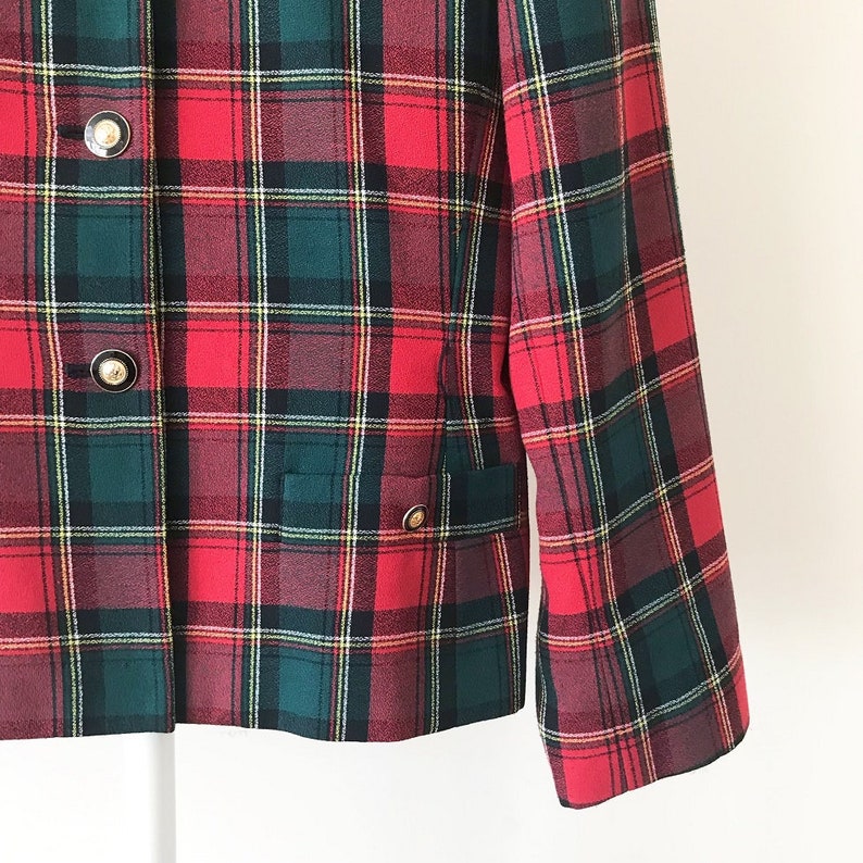 Puede incluir: Una chaqueta de cuadros rojos, verdes y blancos con botones dorados. La chaqueta tiene un bolsillo en el lado izquierdo.