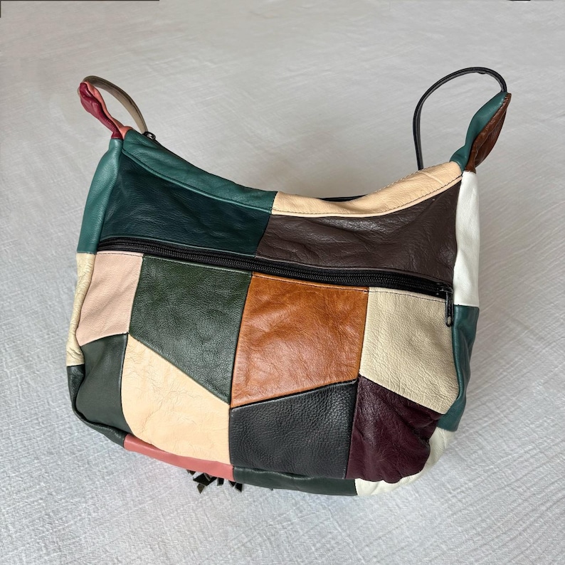 May include: A patchwork leather shoulder bag with a black zipper and handles. The bag features a variety of colors, including green, brown, beige, and black. The bag has a tassel detail on the bottom.