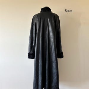 May include: A long black leather coat featuring a high collar and cuffs with a textured black fur-like trim. The coat has a smooth finish, long sleeves, and a straight silhouette. The word Back is printed in black text on the upper right side.