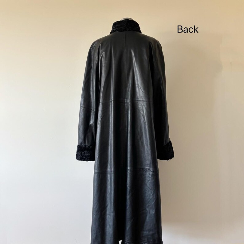 May include: A long black leather coat featuring a high collar and cuffs with a textured black fur-like trim. The coat has a smooth finish, long sleeves, and a straight silhouette. The word Back is printed in black text on the upper right side.