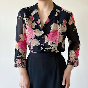 Pode incluir: Uma blusa preta de chiffon com estampa floral com flores rosa e bege. A blusa tem um fecho de botões na frente e mangas compridas com punhos com babados.