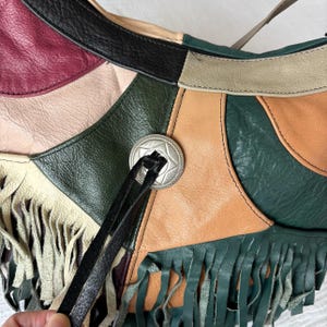 May include: A patchwork leather shoulder bag with a mix of colors including green, tan, burgundy, and cream. The bag features a silver-tone star-shaped button and fringe detailing along the bottom edge. The bag has a long strap.
