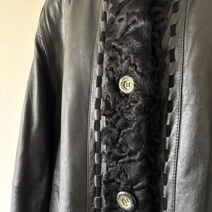 May include: A black leather coat featuring a central panel of textured black curly fur. The coat includes a decorative woven leather trim along the edges of the fur panel and two large, round, metallic buttons with a silver-toned finish.