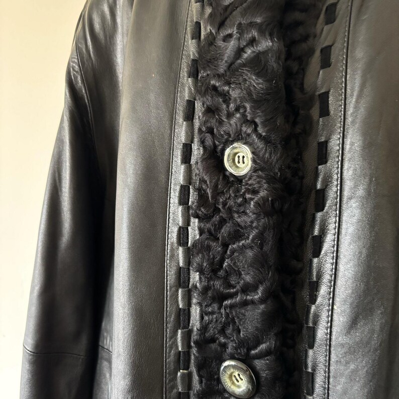 May include: A black leather coat featuring a central panel of textured black curly fur. The coat includes a decorative woven leather trim along the edges of the fur panel and two large, round, metallic buttons with a silver-toned finish.