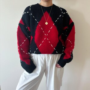 May include: A black, red, and white argyle pattern sweater. The sweater features a crew neck and long sleeves with black cuffs. The argyle pattern is red and black with white stitching. The sweater is made of a thick, textured knit.