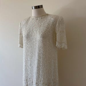 May include: A vintage ivory-colored dress with short sleeves, displayed on a mannequin. The dress is heavily embellished with sequins and beads, creating a textured, shimmering effect. The neckline is adorned with delicate beading.