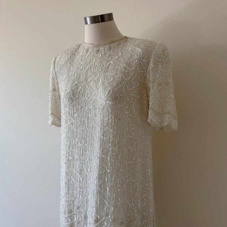 May include: A vintage ivory-colored dress with short sleeves, displayed on a mannequin. The dress is heavily embellished with sequins and beads, creating a textured, shimmering effect. The neckline is adorned with delicate beading.
