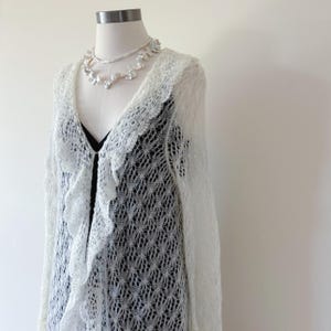 May include: A white, open-front cardigan with a ruffled trim and long sleeves. The cardigan is made of a delicate, open-weave knit. A white floral necklace is also displayed on the mannequin. The cardigan is displayed over a black top.