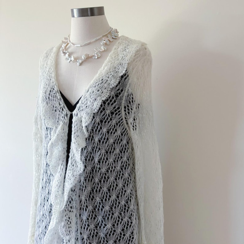 May include: A white, open-front cardigan with a ruffled trim and long sleeves. The cardigan is made of a delicate, open-weave knit. A white floral necklace is also displayed on the mannequin. The cardigan is displayed over a black top.