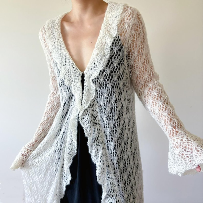 May include: A long, open-front, white knit cardigan with a delicate, openwork design. The cardigan features a V-neckline, ruffled edges, and long sleeves with flared cuffs. The lightweight, airy construction suggests a summer or spring garment.