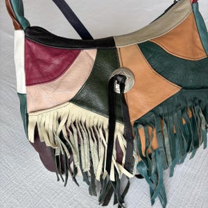 May include: A patchwork leather shoulder bag with a variety of colors including green, tan, burgundy, and black. The bag features a silver-tone concho and fringe detailing along the bottom edge. The strap is black.