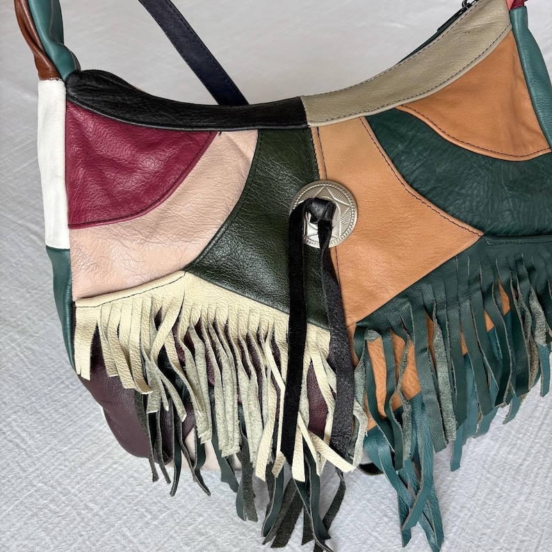 May include: A patchwork leather shoulder bag with a variety of colors including green, tan, burgundy, and black. The bag features a silver-tone concho and fringe detailing along the bottom edge. The strap is black.