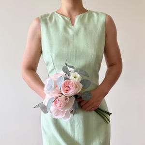 May include: A sleeveless, light green dress with a V-neckline. The dress is paired with a bouquet of pink roses, white flowers, and gray-green leaves. The bouquet is held in front of the dress.