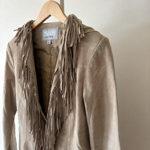 May include: A tan suede jacket with fringe detailing. The jacket features a notched collar and a fringe design along the front and shoulders. The label reads "WILSONS LEATHER MAXIMA". The jacket is hanging on a wooden hanger.