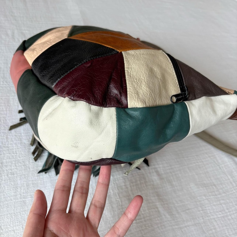 May include: A patchwork leather handbag with a variety of colors including white, green, black, brown, and burgundy. The bag has a zipper closure and fringe detailing. The bag is held by a person.
