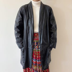 May include: A black leather coat with a wide lapel collar. The coat is long, reaching mid-thigh, and has a relaxed fit. The model is wearing a white turtleneck and a colorful patterned skirt. The coat has a classic, vintage aesthetic.