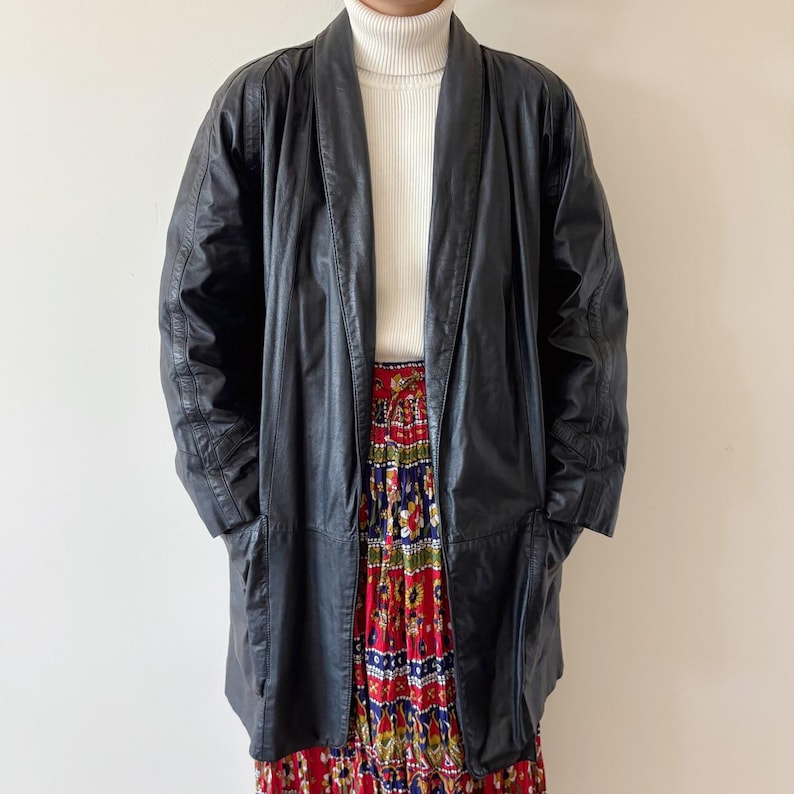 May include: A black leather coat with a wide lapel collar. The coat is long, reaching mid-thigh, and has a relaxed fit. The model is wearing a white turtleneck and a colorful patterned skirt. The coat has a classic, vintage aesthetic.