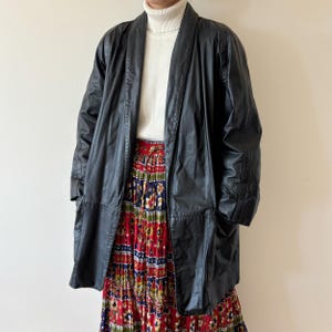 May include: A black leather jacket with a long, open design. The jacket is worn over a white turtleneck and a colorful, patterned skirt with red, blue, and yellow hues. The jacket has a classic, timeless style.