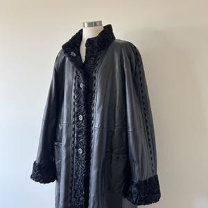 May include: A black leather coat featuring a textured black fur-like collar and cuffs. The coat includes a front button closure, vertical panels of textured trim along the placket, and decorative woven detailing running down the sleeves.