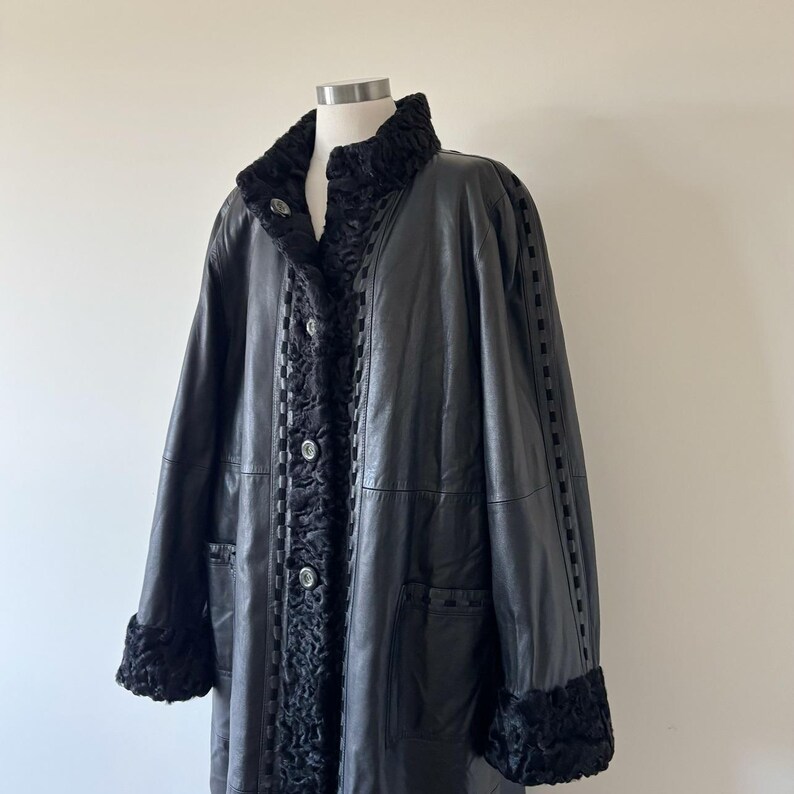 May include: A black leather coat featuring a textured black fur-like collar and cuffs. The coat includes a front button closure, vertical panels of textured trim along the placket, and decorative woven detailing running down the sleeves.