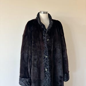 May include: A dark brown coat featuring a textured, curly fur-like trim along the front placket, collar, and sleeve cuffs. The coat has a smooth, solid dark brown body and is fastened with three visible buttons down the center front.