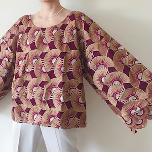 May include: A long-sleeved blouse with a round neckline. The blouse features a repeating pattern of fan-like shapes in burgundy, tan, and silver. The sleeves are wide and the blouse is paired with white pants.