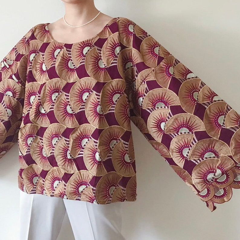 May include: A long-sleeved blouse with a round neckline. The blouse features a repeating pattern of fan-like shapes in burgundy, tan, and silver. The sleeves are wide and the blouse is paired with white pants.